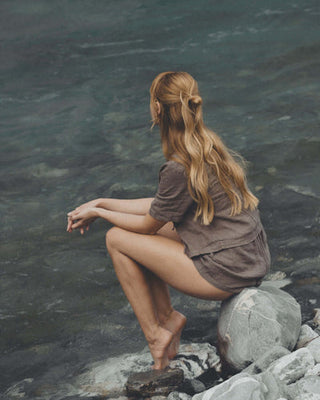 Rothaarige Frau in kurzer brauner Kleidung sitzt auf einem grauen Stein und schaut auf einen Fluss