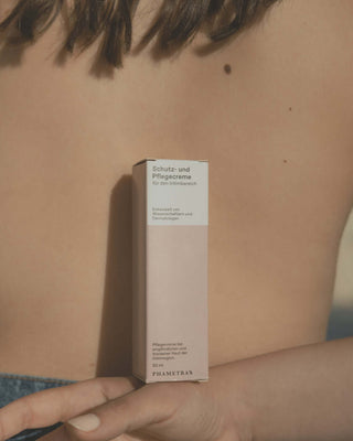 Pinke Schachtel mit Schutz- und Pflegecreme für den Intimbereich wird vor nacktem Rücken gehalten