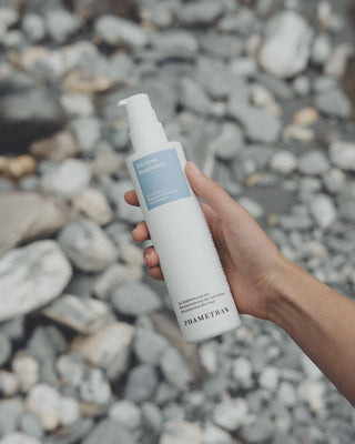 Eine rechte, weibliche Hand hält die 5 % Urea Bodylotion von Phametra mit blauem Etikett in einem weißen Pumpspende vor grauen Steinen