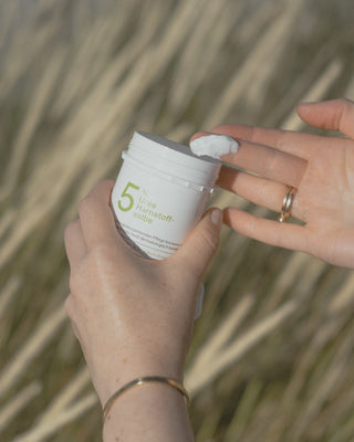 Eine Hand mit goldenen Ringen nimmt Salbe aus einem weißen Plastiktiegel mit 5% Urea Harnstoff-Salbe vor einem Hintergrund mit braunem Farn