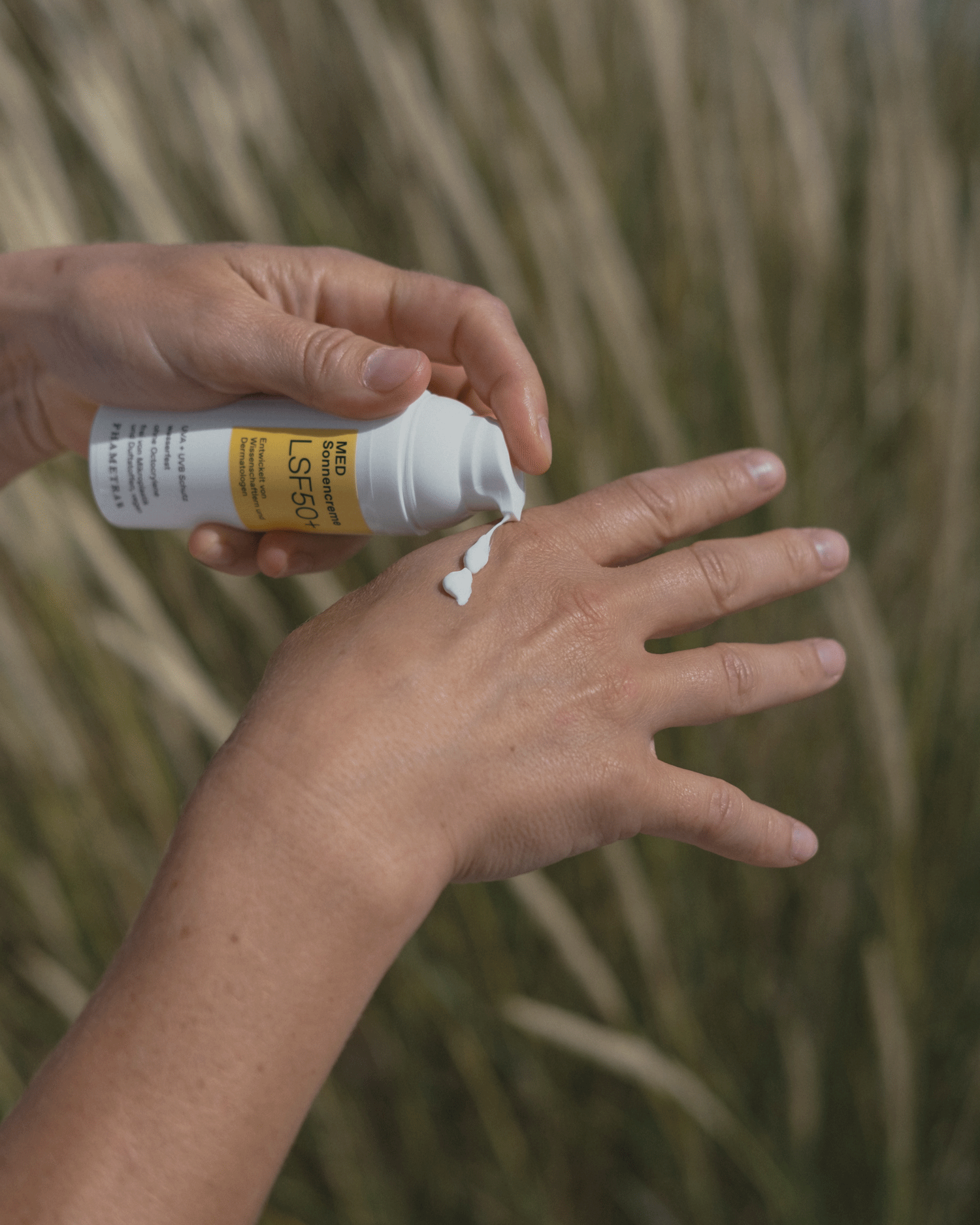Eine Hand gibt Sonnencreme im weiß gelben Spender von Phametra auf einen anderen Handrücken. Im Hintergrund ist Farn zu sehen.