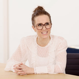Brunette Frau in weißer Bluse mit Dutt und Brille sitzt an hellem Schreibtisch 