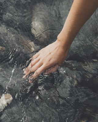 Eine weibliche Hand berührt Wasser in einem Fluss, im Flussbett sind graue Steine zu sehen