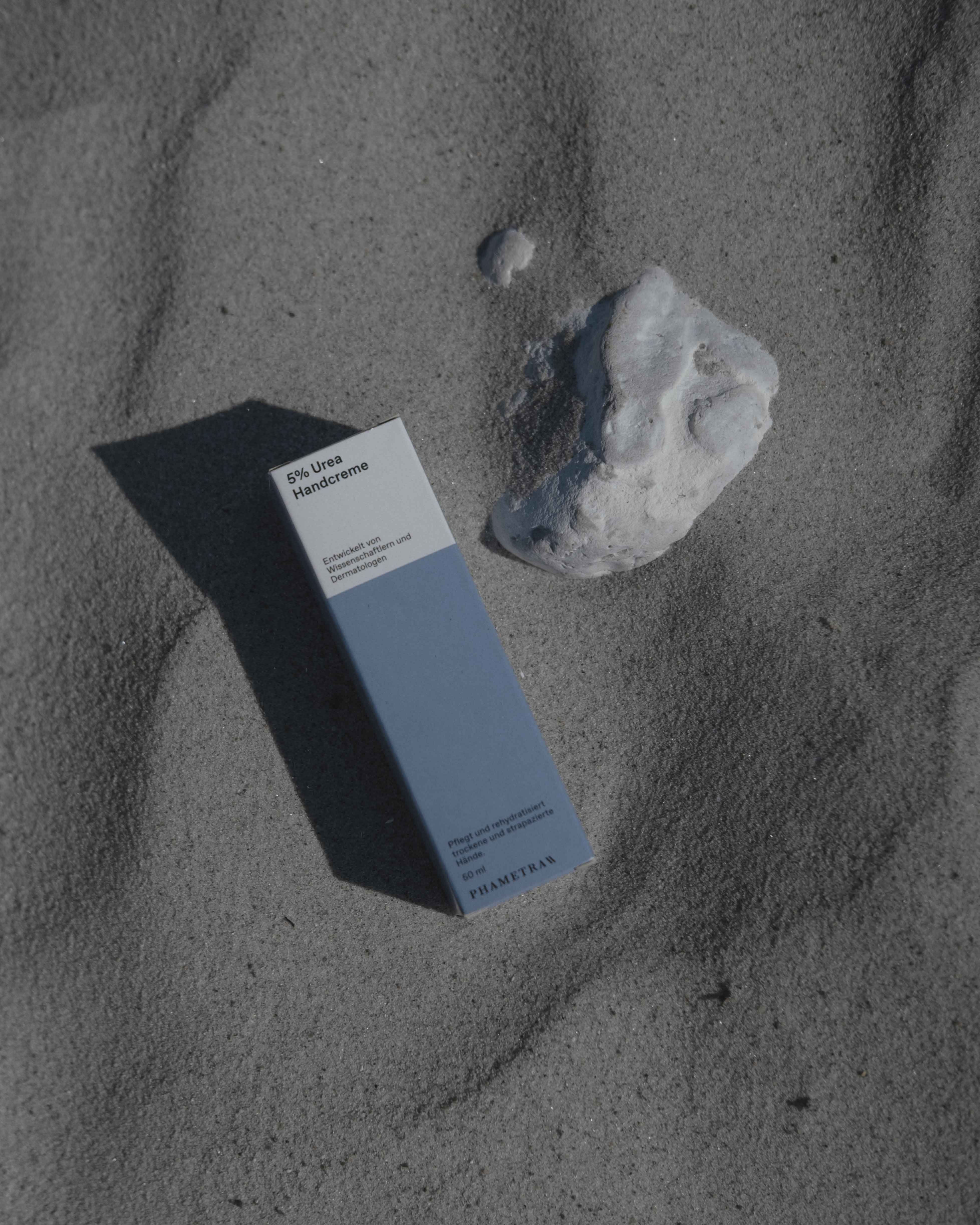 Die blau weiße Umverpackung der Phametra 5% Urea Handcreme liegt im Sand neben einem weißen Stein