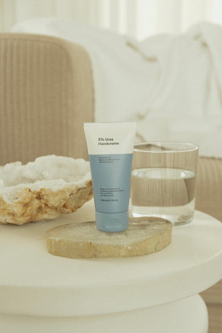 Eine blau weiße Tube mit Phametra Cosmetics 5 % Urea Handcreme steht auf einer beigen Geode auf einem beigen Tisch.