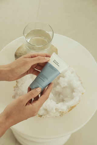 Die 5 % Urea Handcreme in einer blau weißen Tube wird von Frauenhänden vor einen kleinen Couchtisch gehalten. Auf dem Couchtisch steht ein Glas Wasser und eine Geode