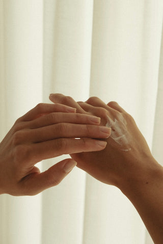 Zwei weibliche Hände cremen sich vor einer weißen Gardine ein