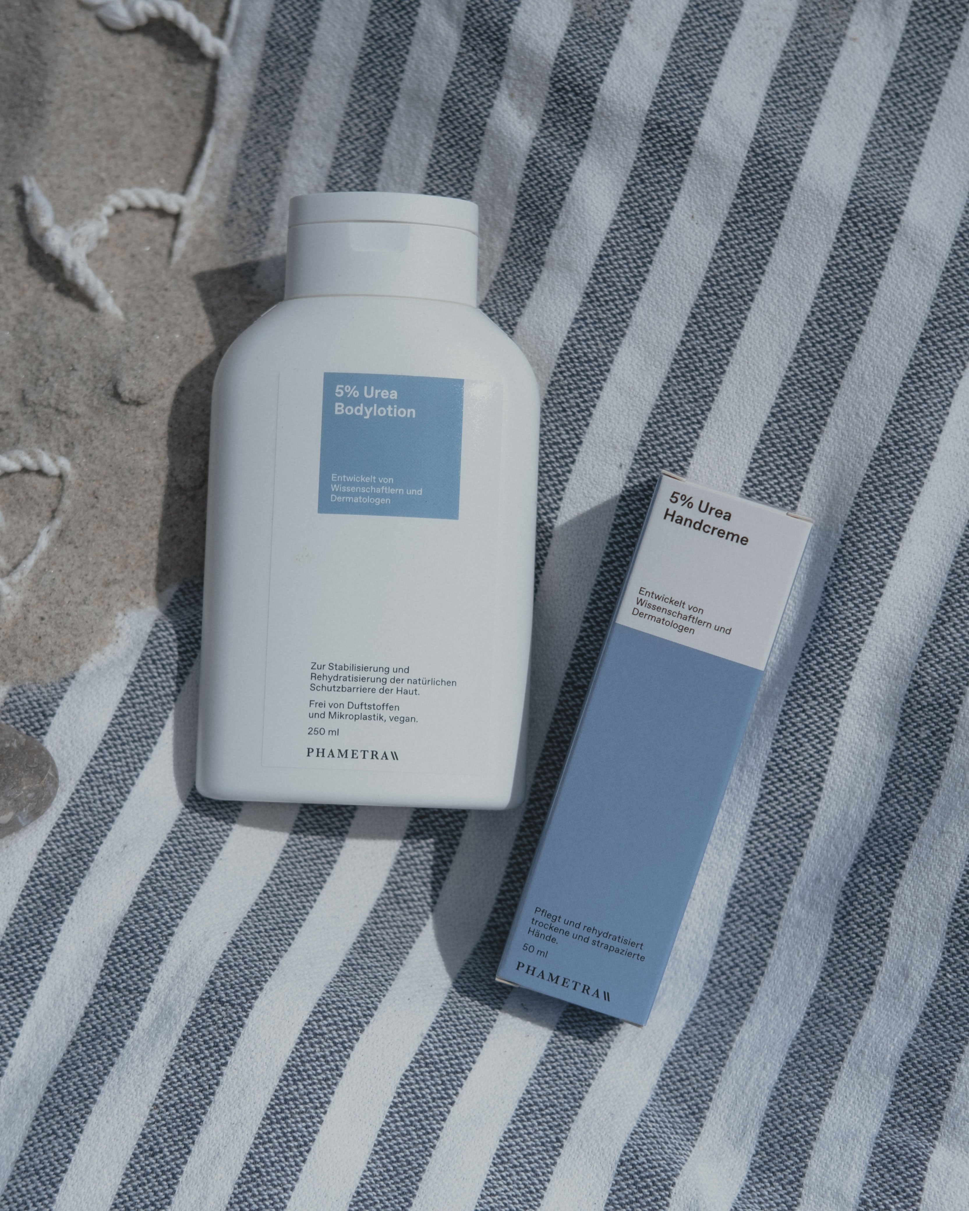 Die weiße Verpackung der Phametra 5% Urea-Bodylotion sowie die blau-weiße Umverpackung der 5% Urea Handcreme von Phametra liegen auf einem blau-weiß gestreifem Handtuch im Sand