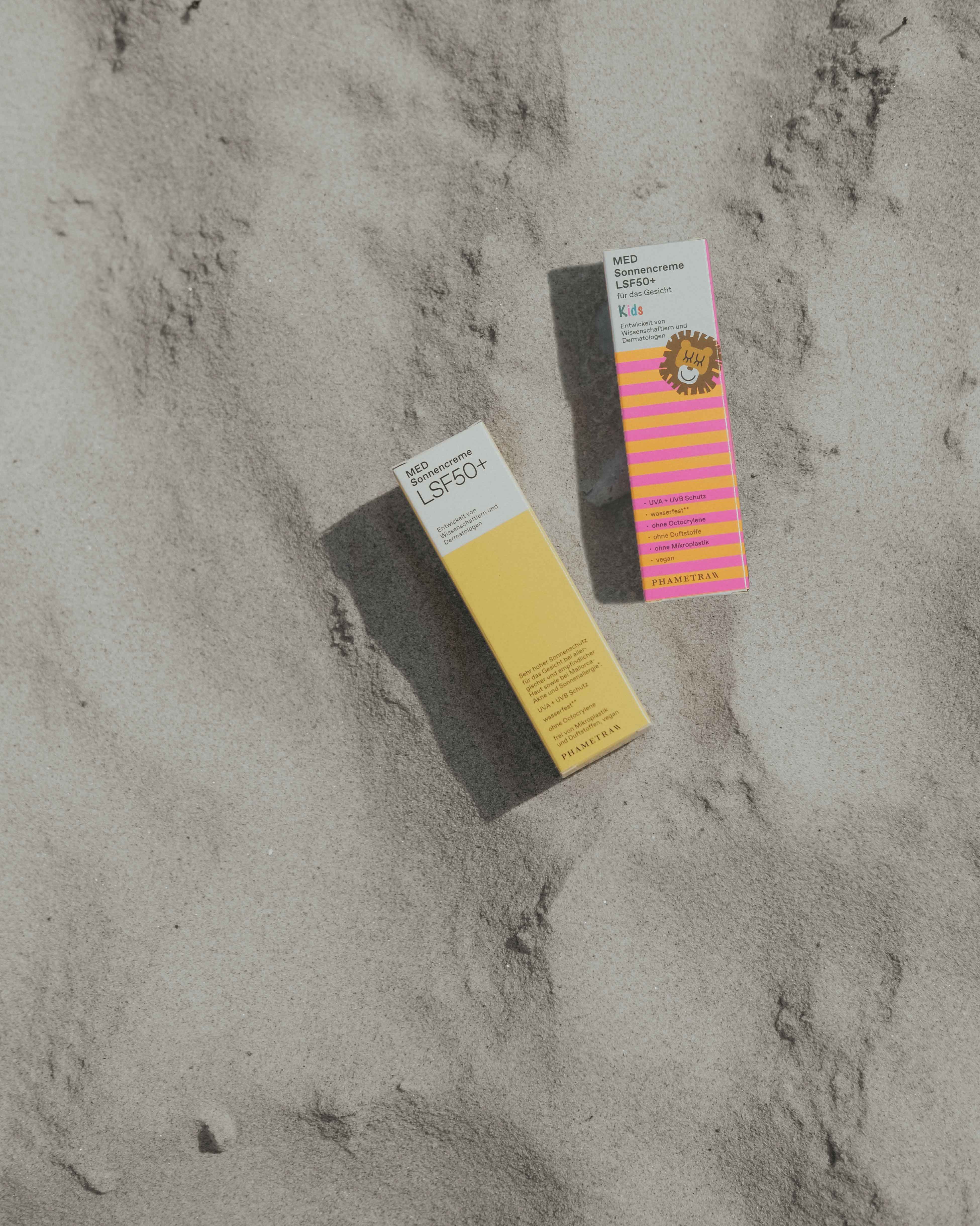 Die gelb-weiße Umverpackung der Phametra Sonnencreme und die weiß-pink-orangene Umverpackung der Phametra Sonnencreme Kids liegen im Sand