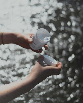 Eine Hand mit goldenen Ringen nimmt Salbe aus einem weißen Plastiktiegel mit 10% Urea Harnstoff-Salbe vor dunklem Wasser