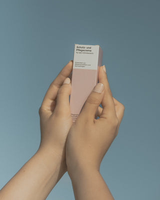 Zwei weibliche Hände halten eine rosane Verpackung der Phametra Schutz- und Pflegecreme für den Intimbereich vor blauem Hintergrund