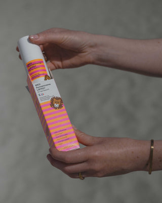 Eine Hand holt am Strand einen orange-pink-weißen Spender mit Phametra Sonnencreme KIDS aus seiner bunten Umverpackung