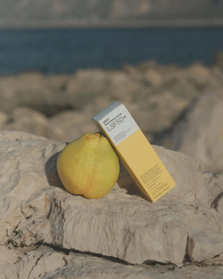 Eine weiß-gelbe Schachtel mit Phametra Sonnencreme ist an eine gelbgrüne Birne gelehnt. Beide stehen auf eine Felsformation. Im Hintergrund ist Wasser zu sehen.