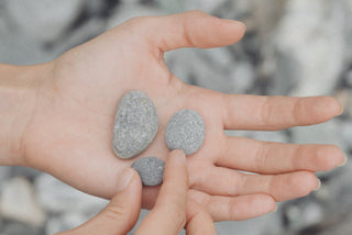 Auf einer weiblichen linken Hand liegen drei graue, kleine und runde Steine, wobei der kleinste Stein von den Fingern der rechten Hand berührt wird. Im Hintergrund sind ebenfalls graue Steine zu sehen.