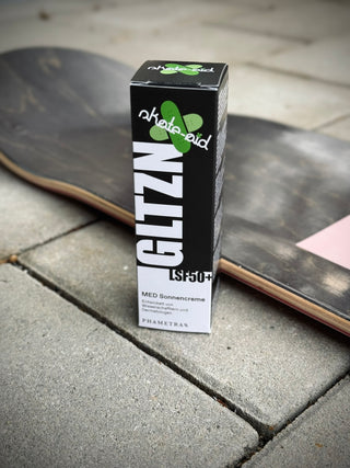 Eine schwarze Verpackung mit GLTZN-Sonnencreme mit Skate-Aid Aufkleber steht auf einem gepflasterten Boden mit Skateboard im Hintergrund